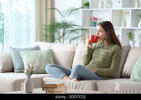 Donna in verde che beve caffè da una tazza rossa che guarda lontano seduto su un divano di casa Foto Stock