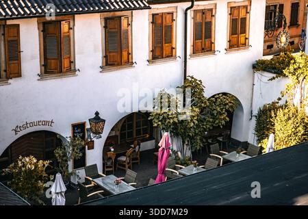 Un affascinante cortile ristorante dall'atmosfera europea, caratterizzato da edifici bianchi, persiane in legno e una varietà di tavoli e sedie. Foto Stock