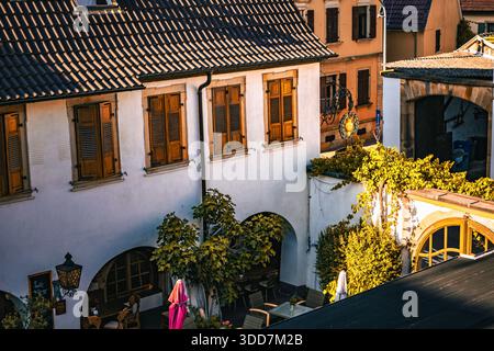Una pittoresca scena di una piazza cittadina europea, che mostra architettura tradizionale, caffetterie all'aperto e una vibrante vegetazione. Foto Stock