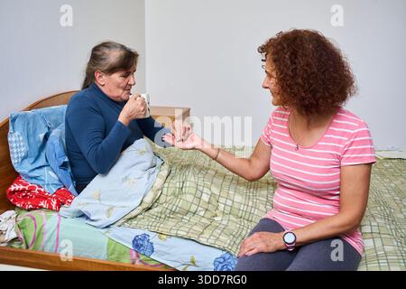Assistente che dà pillole a una donna anziana malata che tiene una tazza di tè mentre è seduta a letto Foto Stock