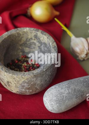 Mortaio e pestello con grani di pepe misti, aglio e cipolla su tessuto rosso Foto Stock