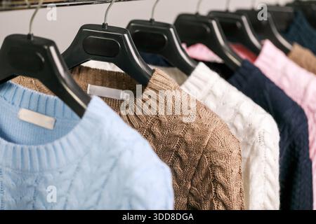 Fila di maglioni lavorati a maglia nelle tonalità blu, marrone, rosa, bianco e navy, disposti in modo ordinato su appendiabiti neri in un negozio di abbigliamento, mettendo in risalto i loro motivi a maglia a cavo e l'accogliente fascino invernale Foto Stock