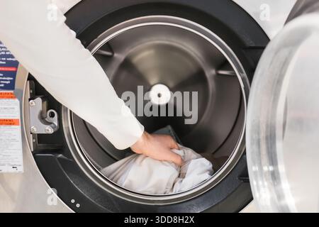 Primo piano della mano maschile che mette abiti bianchi nel tamburo della lavatrice industriale aperto presso la lavanderia automatica. Foto di alta qualità Foto Stock