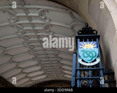 Manchester, Regno Unito - 06 dicembre 2025: Stemma decorativo con sole e emblema intrecciato su un cancello di ferro sotto uno storico arco di Manchester. Foto Stock