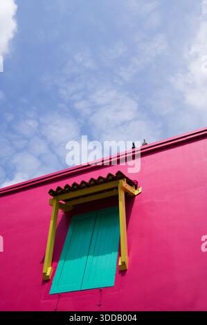 Colpo ritagliato di una finestra quadrata su un muro rosa, contro un cielo blu chiaro. Due piccioni seduti sul tetto. Foto Stock
