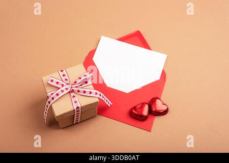 Busta rossa con carta bianca, confezione regalo e caramelle a forma di cuore su sfondo beige. Il giorno di San Valentino. Vista dall'alto, posizione piatta, mockup. Foto Stock