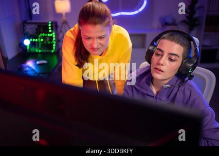 Due donne che indossano le cuffie che giocano insieme a un videogioco in una sala giochi colorata. Sono in possesso di controller di gioco, interagiscono con lo schermo e si divertono in una sessione multigiocatore entusiasmante || Model rilasciato Foto Stock
