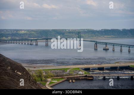 Un lungo ponte sospeso in acciaio verde attraversa un ampio fiume con colline boscose sullo sfondo e un ponte stradale più piccolo in primo piano. Quebec City Foto Stock