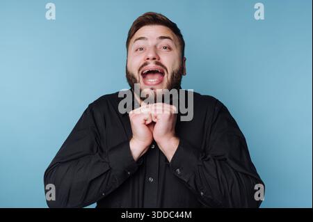 Uomo eccitato in camicia nera che esprime gioia su sfondo blu. Foto Stock