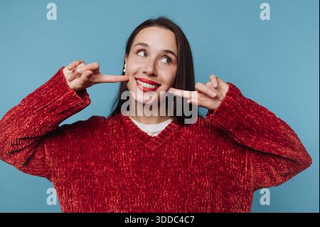 Donna in maglione rosso che sorride e punta le guance Foto Stock