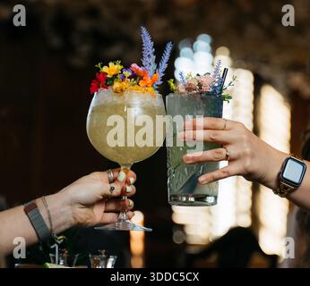Due persone reggono bicchieri di bevande, uno dei quali ha una decorazione floreale. La scena è ambientata in un bar o ristorante, e le bevande sono probabilmente coc Foto Stock