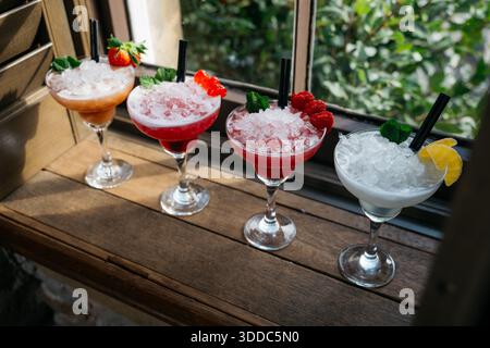 Quattro bevande di diversi colori in bicchieri martini con cannucce. Le bevande sono guarnite con frutta e ghiaccio Foto Stock