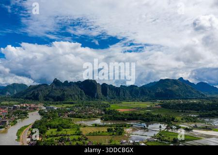 Ampia vista aerea di Vang Vieng, Laos, con spettacolari scogliere calcaree che si innalzano dietro un mosaico di risaie e il fiume Nam Song che si snoda attraverso la lussureggiante valle. Le ampie nuvole e il vivace cielo blu incorniciano l'impressionante paesaggio carsico durante la stagione delle piogge. Foto Stock