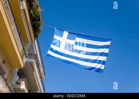Grande bandiera greca sospesa tra i balconi residenziali di Arta, Grecia, adagiata su un cielo azzurro. La luce solare nitida mette in risalto il tessuto e l'architettura urbana, creando un suggestivo paesaggio urbano con linee geometriche. Foto Stock