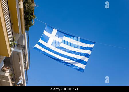 Grande bandiera greca si estendeva attraverso lo spazio tra i balconi residenziali di Arta, in Grecia, adagiato su un cielo blu vivido. La luce del sole mette in risalto il tessuto fresco e l'architettura urbana, creando una suggestiva scena cittadina. Foto Stock