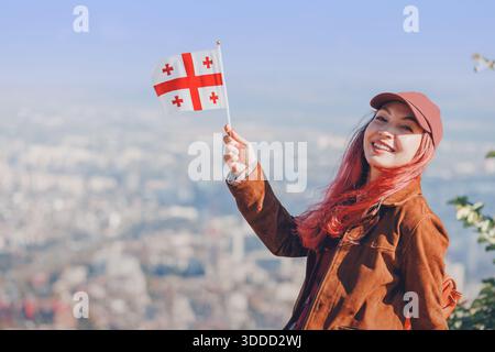 Giovane donna con i capelli rosa sorridente, con la bandiera georgiana in alto su uno sfondo urbano sfocato. Concetti di patriottismo e viaggio Foto Stock