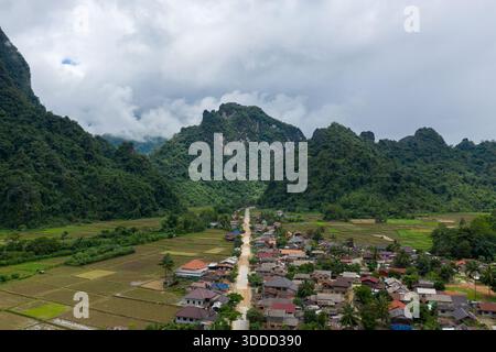 Un villaggio rurale si trova annidato alla base di ripide colline carsiche boscose con un'unica strada che corre attraverso campi di riso patchwork nella campagna tra Vang Vieng e Phou Khoun, Laos. La scena presenta una vegetazione vivace, terreni agricoli geometrici e spettacolari nuvole appese in basso sul lussureggiante paesaggio. Foto Stock