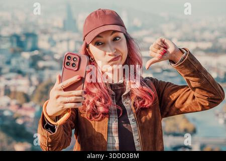 Giovane donna con i capelli rosa che mostra disapprovazione, con il pollice abbassato, mentre scatta un selfie con lo smartphone in città Foto Stock