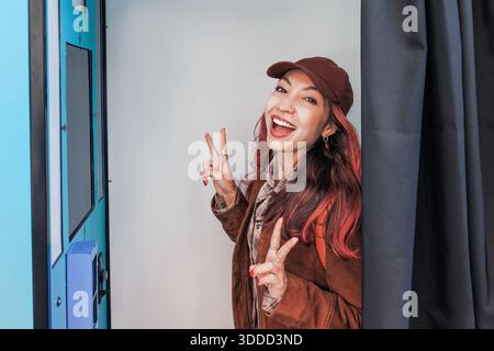 Giovane donna sorridente, che fa segnali di pace e mostra un'espressione vibrante all'interno di uno stand fotografico per ricordi divertenti Foto Stock