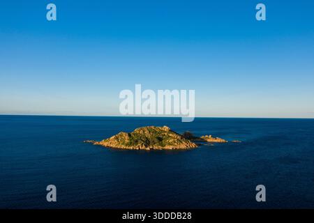 Una piccola isola aspra coperta da vegetazione verde si trova da sola nel mare calmo e blu sotto un vasto cielo limpido vicino a Lotzorai, in Sardegna. La calda luce del sole della sera mette in risalto la consistenza delle rocce e del fogliame. Foto Stock