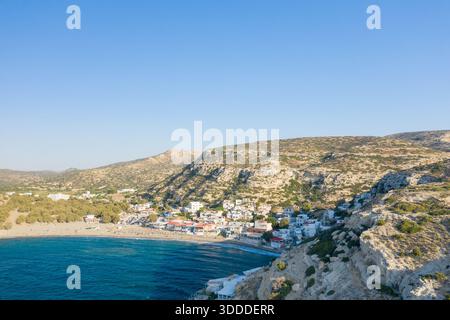 Il villaggio di Matala sorge tra una baia sabbiosa e colline calcaree illuminate dal sole sulla costa meridionale di Creta. L'acqua blu cristallina contrasta con le formazioni rocciose testurizzate e gli edifici imbiancati sotto un cielo senza nuvole. Foto Stock