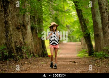 Il bambino con zaino e cappello cammina attraverso la foresta, guardando in avanti. Escursione nella natura in estate. Momenti di gioia all'aperto durante le vacanze e le vacanze attive. Foto Stock
