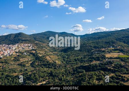 Vista aerea del villaggio di Orgosolo ai margini di colline densamente boscose e campi di patchwork sotto un cielo azzurro luminoso con nuvole sparse in Sardegna. Foto Stock