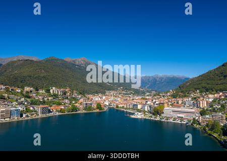 Panorama ad alto angolo di Omegna sul Lago d'Orta, nell'Italia settentrionale, che mostra moderni edifici sul lungomare, un centro città densamente costruito e colline boscose con aspre vette alpine in lontananza sotto un cielo blu vivido. La tranquilla superficie del lago e la vegetazione lussureggiante creano un paesaggio fresco e invitante. Foto Stock