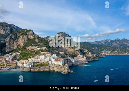 Un'ampia prospettiva aerea cattura gli edifici imbiancati di Amalfi, Italia, annidati lungo le scogliere spettacolari sopra il profondo blu del Mar Mediterraneo. Aspre montagne, colline terrazzate e una barca a vela solitaria completano la tranquilla scena costiera sotto un cielo limpido. Foto Stock