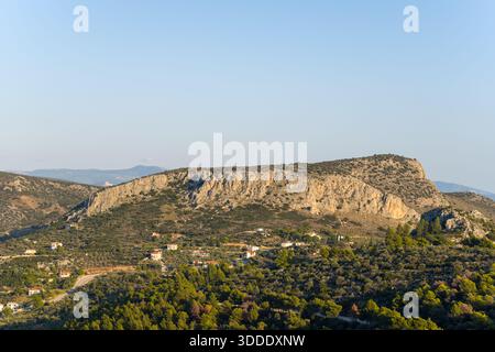 Ampia vista panoramica di un'aspra cresta calcarea che si innalza sopra un mosaico di oliveti e case rurali sparse nella campagna greca. La calda luce del sole della sera proietta ombre delicate attraverso i pendii rocciosi e la vegetazione verde. Foto Stock