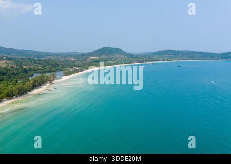 L'ampia vista aerea mostra il delicato arco di Long Set Beach delimitato da acque turchesi cristalline e da una lussureggiante foresta verde sull'isola di Koh Rong, Cambogia. Colline ondulate e barche sparse completano questa tranquilla costa tropicale. Foto Stock