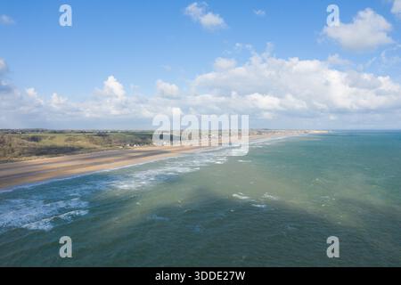 Un'ampia vista aerea mostra l'ampia costa sabbiosa di Omaha Beach delimitata da colline verdi ondulate e case sparse, con dolci onde che incontrano la costa sotto un cielo luminoso pieno di nuvole sparse. Foto Stock