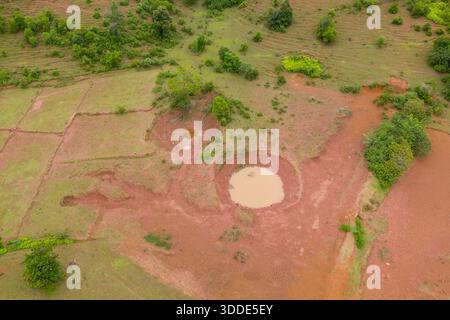 Prospettiva droni di stagni fangosi circolari e praterie verdi discontinue sull'altopiano di Plaine des Jarres in Laos. Il paesaggio presenta terra rossastra, arbusti sparsi e sottili texture naturali sotto la luce del giorno. Foto Stock
