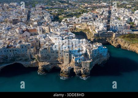 Ampia vista aerea di Polignano a Mare in Puglia, Italia, mostrando edifici storici imbiancati raggruppati lungo le suggestive scogliere calcaree sopra il blu profondo del mare Adriatico. Il paesaggio urbano costiero contrasta le aspre formazioni rocciose con la densa architettura mediterranea sotto la luce del sole. Foto Stock