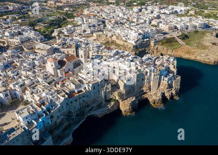 La prospettiva aerea rivela edifici imbiancati densamente pieni in cima a spettacolari scogliere calcaree che si affacciano sul blu profondo del Mar Adriatico a Polignano a Mare, Puglia, Italia. La storica città costiera contrasta le aspre formazioni rocciose con l'architettura mediterranea sotto la luce del sole. Foto Stock