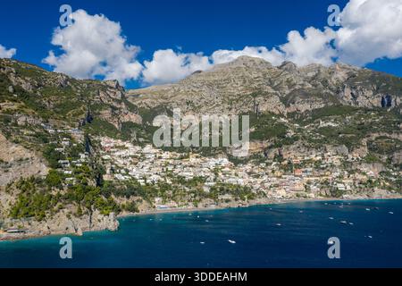 Un'ampia prospettiva aerea rivela il villaggio costiero di Positano, in Italia, annidato tra ripide scogliere calcaree e le acque blu profonde della Costiera Amalfitana. Le spettacolari montagne si innalzano sopra edifici terrazzati, con barche sparse che punteggiano il vibrante mare Mediterraneo sotto un cielo luminoso. Foto Stock