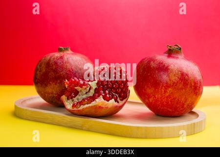 Due melograni interi e uno tagliato aperto con semi rossi sono su un vassoio di legno Lo sfondo ha colori rosso e giallo luminosi. Questa scena si svolge io Foto Stock