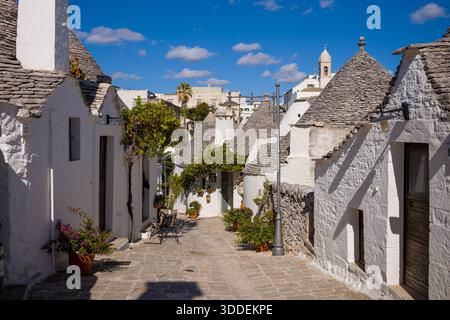 Affascinante strada stretta fiancheggiata da tradizionali case a trullo dipinte di bianco e tetti in pietra ad Alberobello, in Puglia, in Italia. Le piante in vaso, le consistenze rustiche e il cielo azzurro creano una vibrante atmosfera italiana meridionale. Foto Stock