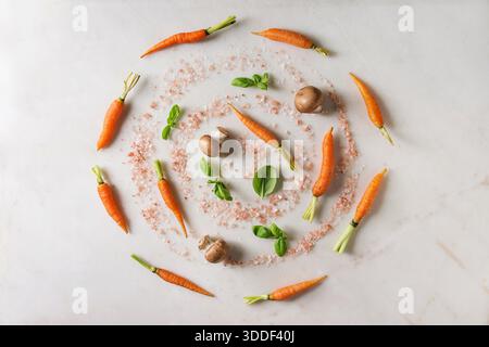 Erbe fresche di basilico con piccole carote, sale rosa e funghi champignon a spirale su fondo in marmo bianco. Piatto, spazio. Concetto di cucina, background gastronomico. Foto Stock