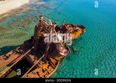 Prospettiva aerea del relitto arrugginito di Dimitrios parzialmente sommerso in acque turchesi chiare e vibranti vicino a una costa sabbiosa vicino a Gythio, Grecia. La luce del sole illumina il metallo corroso e rivela formazioni rocciose sottomarine. Foto Stock