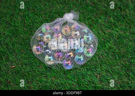 Biglie in vetro trasparente con rivestimento iridescente racchiuse in una borsa a rete bianca, su una vivace erba verde artificiale Foto Stock