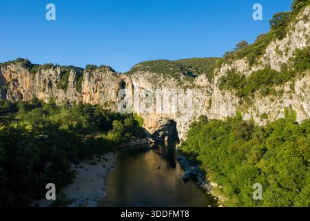 L'alta prospettiva rivela spettacolari scogliere calcaree e una fitta foresta verde che fiancheggia un tortuoso fiume a Pont d'Arc ad Ardeche, sotto un vivace cielo blu. Le texture robuste e le ombre profonde creano un paesaggio naturale sorprendente. Foto Stock