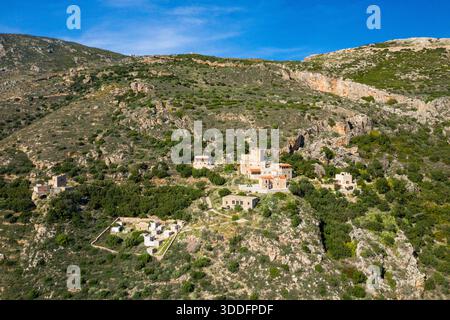 Prospettiva aerea delle tradizionali case in pietra annidate tra pendii rocciosi e vegetazione lussureggiante su una collina illuminata dal sole a mani, in Grecia, sotto un cielo azzurro. Foto Stock