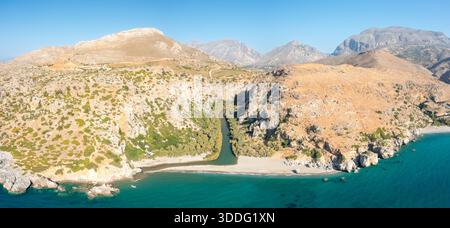 Ampia vista aerea del fiume Preveli che scorre attraverso una lussureggiante gola fiancheggiata da palme per incontrare la spiaggia sabbiosa e il turchese Mar Mediterraneo, circondato da montagne aride e aspre scogliere sotto un cielo azzurro. Foto Stock