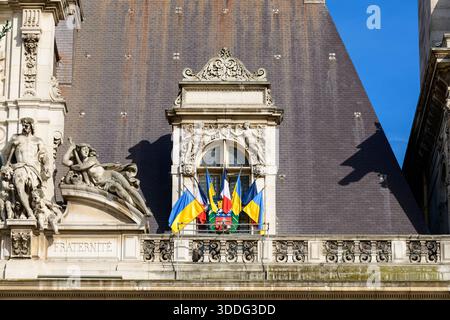 Bandiere ucraine e francesi sono esposte sopra un balcone ornato con sculture in pietra classiche e intricate incisioni sullo storico Hotel de Ville di Parigi. La luce del sole mette in risalto la facciata dettagliata e gli elementi architettonici decorativi. Foto Stock