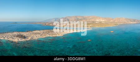 Ampia vista aerea panoramica della spiaggia di Elafonisi a Creta, in Grecia, con un mare turchese vivace, una costa rocciosa e colline aride illuminate dal sole sotto un cielo azzurro. Il paesaggio sembra ampio, naturale e sereno. Foto Stock