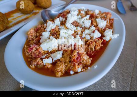 Primo piano di un piatto greco rustico con uova strapazzate cotte con pomodori e ricoperte di feta sbriciolata, servite su un piatto bianco. I colori vivaci e la consistenza abbondante evocano un pasto mediterraneo fatto in casa. Foto Stock