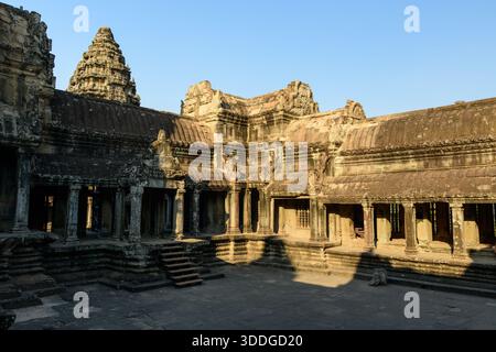 La calda luce del sole del tardo pomeriggio proietta ombre attraverso le gallerie di arenaria e il cortile a gradini sulla cima di Angkor VAT, Cambogia. Le intricate tetti e gli antichi dettagli architettonici Khmer si distinguono sotto un cielo azzurro. Foto Stock