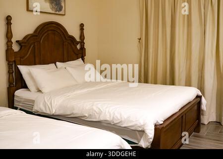 Camera da letto ordinata con due letti, biancheria bianca, testiera in legno, tende beige e arte astratta delle pareti in un ambiente caldo e invitante. Foto Stock