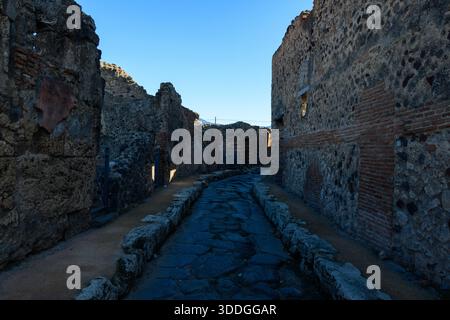 Uno stretto e antico vicolo romano fiancheggiato da mura in pietra vulcanica e mattoni a Pompei, Italia. Ombre profonde e macchie di luce solare mettono in risalto la grezza muratura e le trame storiche sotto un cielo blu limpido. Foto Stock